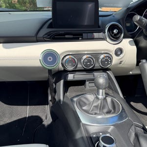 May include: Interior view of a car dashboard. The dashboard is light-colored with a black infotainment screen. The center console features climate control knobs, a gear shift, and a control dial. A circular object with a green and black design is attached to the dashboard.
