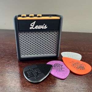 May include: A small black and orange electric guitar amplifier with the word "Lewis" printed on the front. The amplifier is sitting on a wooden surface with four guitar picks in front of it.