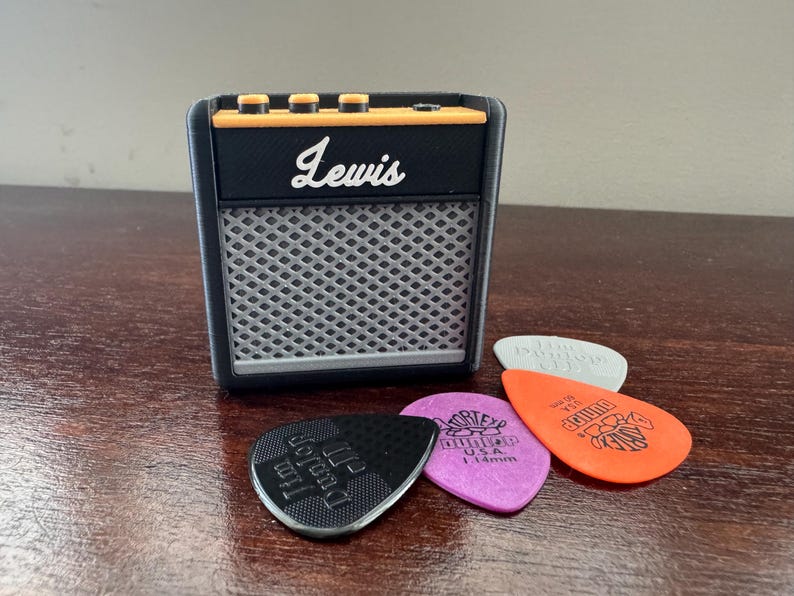 May include: A small black and orange electric guitar amplifier with the word "Lewis" printed on the front. The amplifier is sitting on a wooden surface with four guitar picks in front of it.