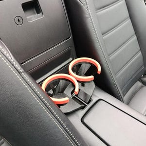 May include: A car interior with black leather seats and a center console. Two pairs of sunglasses with orange frames rest in a black cup holder. The console has a storage compartment and a keyhole.