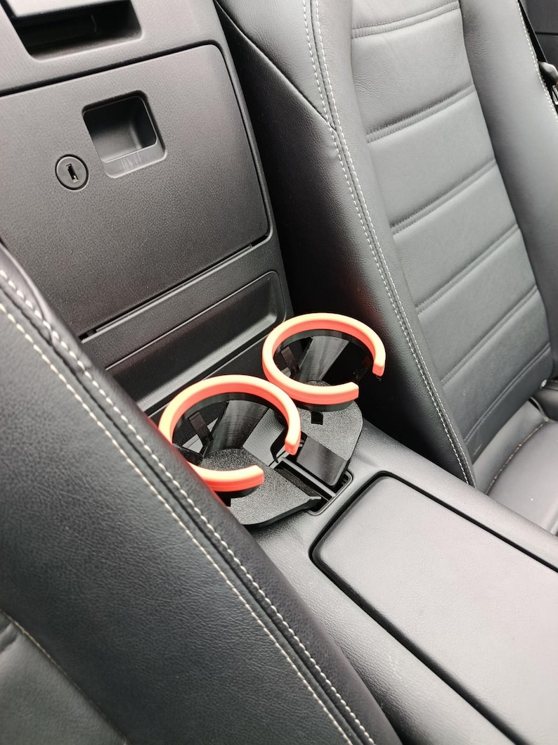 May include: A car interior with black leather seats and a center console. Two pairs of sunglasses with orange frames rest in a black cup holder. The console has a storage compartment and a keyhole.