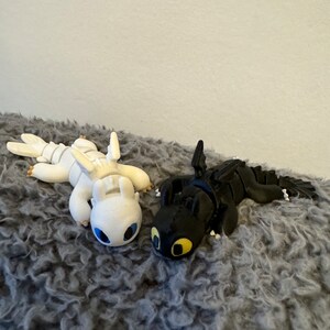 May include: Two small dragon figurines, one white with blue eyes and one black with yellow eyes, lying on a gray fuzzy surface.