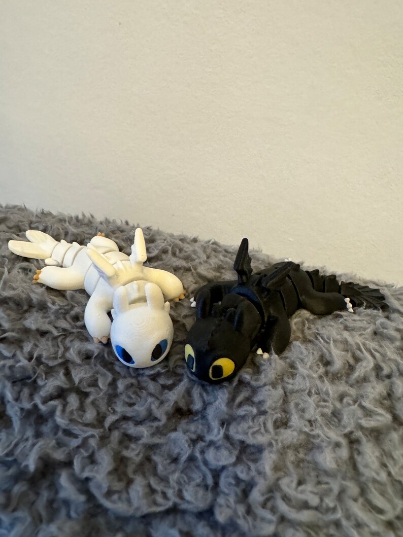 May include: Two small dragon figurines, one white with blue eyes and one black with yellow eyes, lying on a gray fuzzy surface.