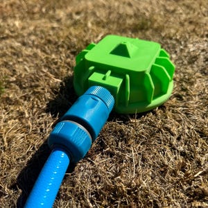 May include: A bright green sprinkler head with a blue hose connection. The sprinkler head has a square top and a circular base with fins. The blue hose is connected to the sprinkler head and is lying on dry grass.
