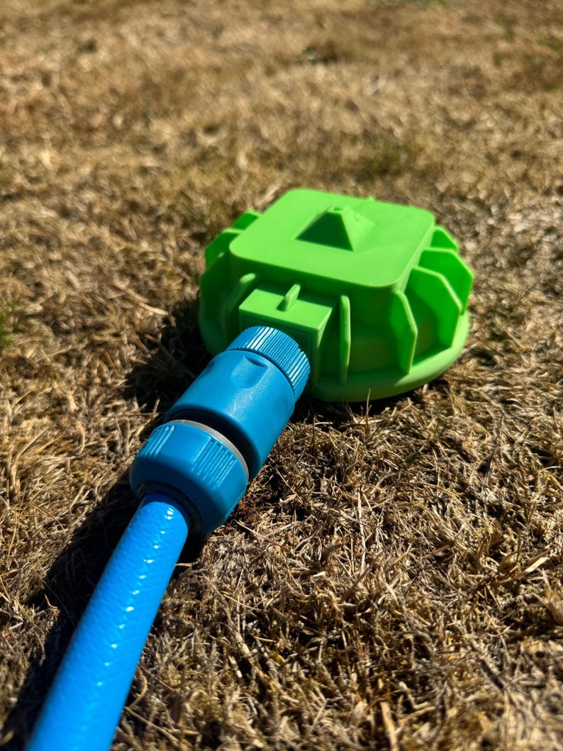 May include: A bright green sprinkler head with a blue hose connection. The sprinkler head has a square top and a circular base with fins. The blue hose is connected to the sprinkler head and is lying on dry grass.