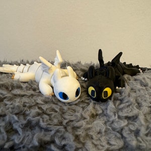 May include: Two 3D printed dragon figurines, one white and one black, lying on a gray fuzzy surface. The white dragon has blue eyes and the black dragon has yellow eyes.