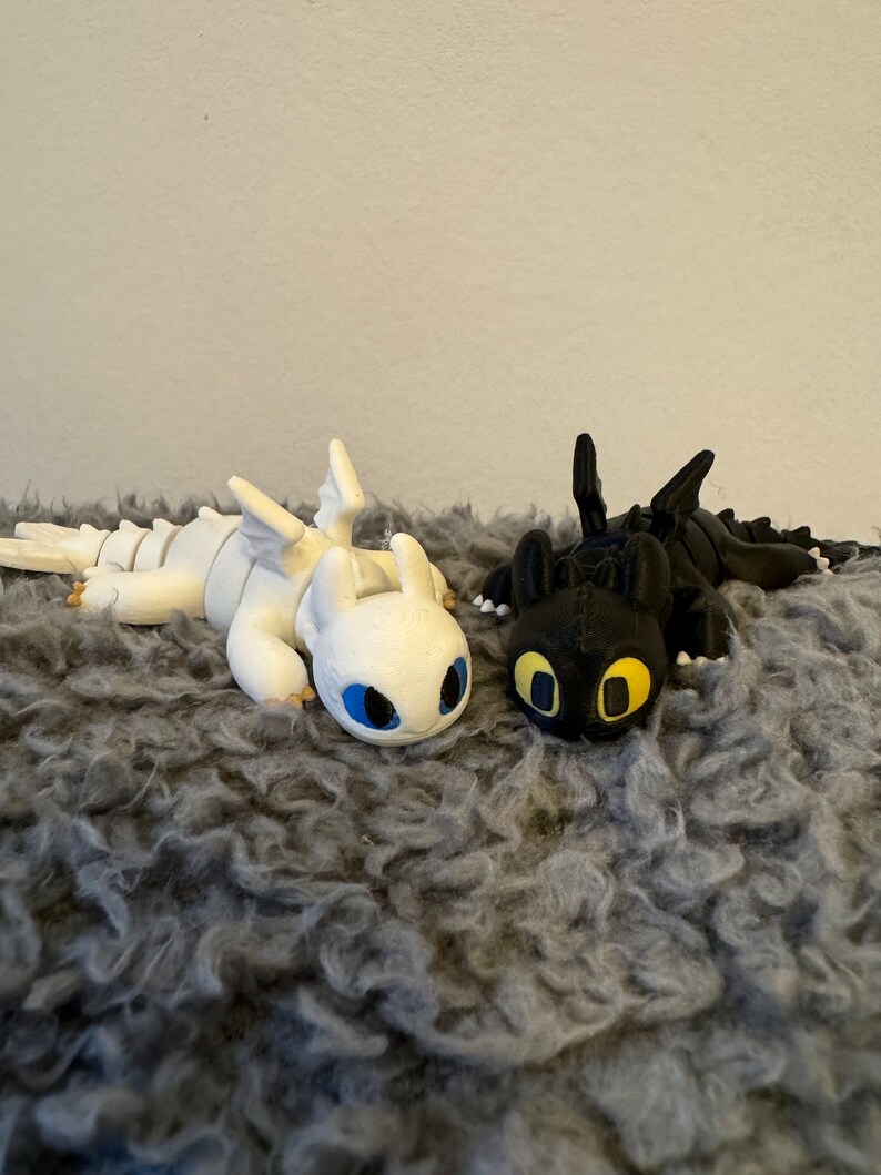 May include: Two 3D printed dragon figurines, one white and one black, lying on a gray fuzzy surface. The white dragon has blue eyes and the black dragon has yellow eyes.