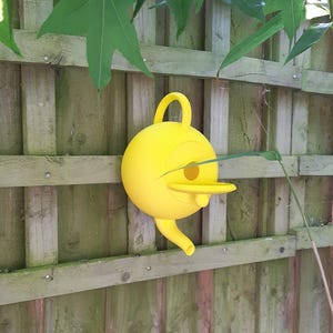 May include: A bright yellow birdhouse with a round body, a beak-like entrance, and a curved handle. The birdhouse is mounted on a weathered wooden fence, with green leaves and branches in the background.