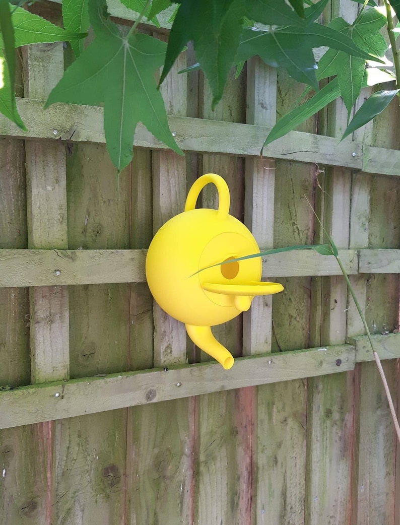 May include: A bright yellow birdhouse with a round body, a beak-like entrance, and a curved handle. The birdhouse is mounted on a weathered wooden fence, with green leaves and branches in the background.