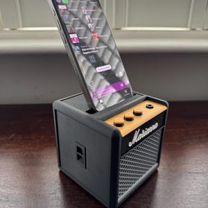 May include: A black and orange miniature guitar amplifier with a smartphone resting in the top slot. The phone screen shows a music streaming app with the text "Continue watching" visible. The amplifier has the word "Marshall" printed on the front.