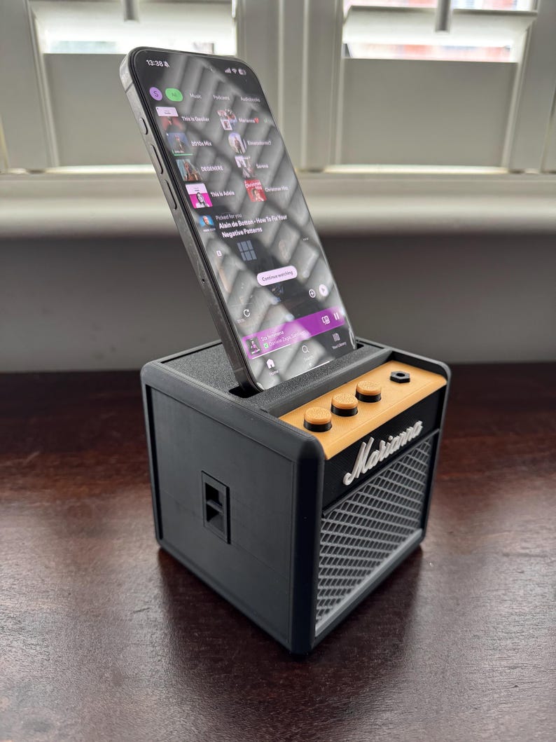 May include: A black and orange miniature guitar amplifier with a smartphone resting in the top slot. The phone screen shows a music streaming app with the text "Continue watching" visible. The amplifier has the word "Marshall" printed on the front.