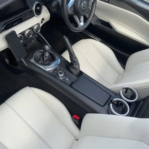 Puede incluir: Vista interior de un coche descapotable con asientos de cuero blanco y un salpicadero negro. El volante es negro con detalles plateados, y la consola central tiene una palanca de cambios y portavasos. Un teléfono está montado en el salpicadero.