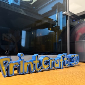 May include: A blue and yellow 3D printed sign that reads "Printcraft 3D".