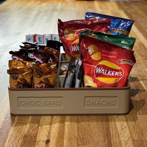May include: A beige, divided snack organizer labeled "CHOC BARS" and "SNACKS" filled with various packaged snacks. The snacks include chocolate bars and bags of potato chips in red, blue, and green packaging.