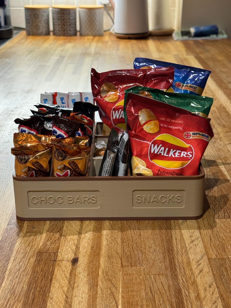 May include: A beige, divided snack organizer labeled "CHOC BARS" and "SNACKS" filled with various packaged snacks. The snacks include chocolate bars and bags of potato chips in red, blue, and green packaging.