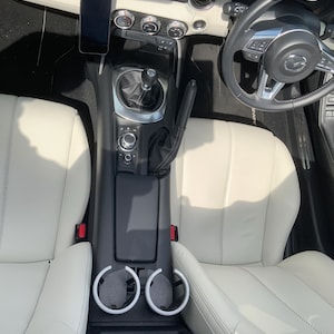 May include: Interior view of a car with white leather seats, a black dashboard, and a black steering wheel. A smartphone is mounted on the dashboard. The center console has cup holders and a gear shift.