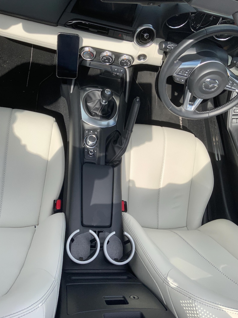 May include: Interior view of a car with white leather seats, a black dashboard, and a black steering wheel. A smartphone is mounted on the dashboard. The center console has cup holders and a gear shift.