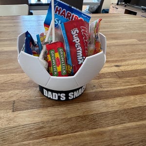 Op de afbeelding: Een witte, 3D-geprinte container in de vorm van een voetbal, gevuld met snoep en snacks. De basis heeft de tekst "DAD'S SNACKS" in het zwart. De container staat op een houten oppervlak.