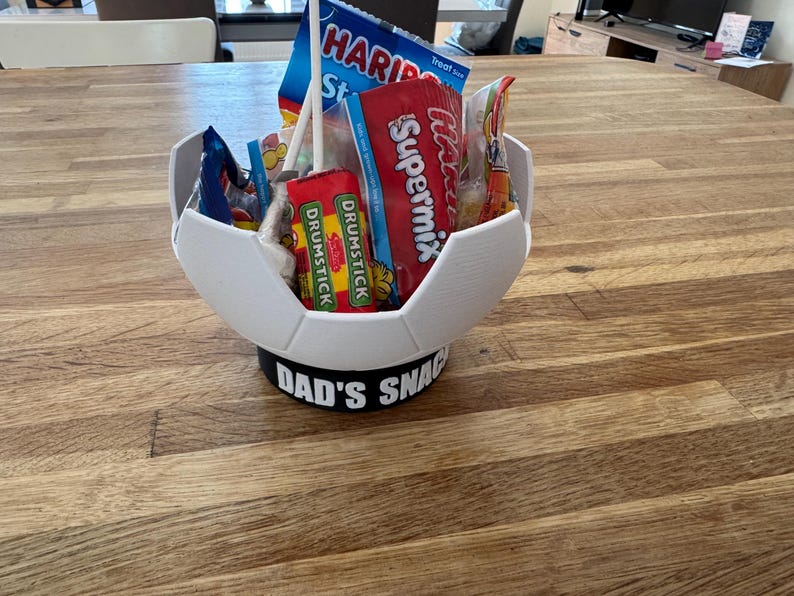 Op de afbeelding: Een witte, 3D-geprinte container in de vorm van een voetbal, gevuld met snoep en snacks. De basis heeft de tekst "DAD'S SNACKS" in het zwart. De container staat op een houten oppervlak.