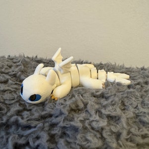 May include: White articulated dragon figurine with blue eyes, lying on a gray fuzzy surface.