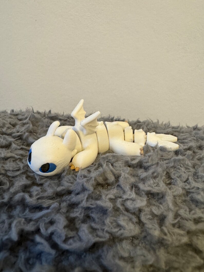 May include: White articulated dragon figurine with blue eyes, lying on a gray fuzzy surface.