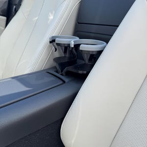 May include: Interior view of a car featuring a black cup holder with two gray inserts, positioned between white leather seats. The seats have a perforated design and are stitched with white thread. A red button is visible.