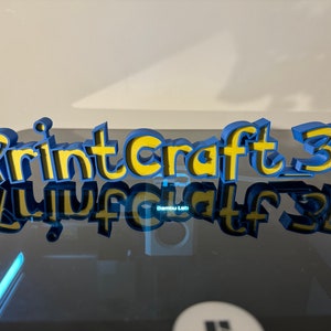 May include: A 3D printed sign that reads "Printcraft 3D" in blue and yellow letters. The sign is sitting on a black surface and is reflected in the surface.