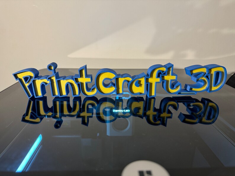 May include: A 3D printed sign that reads "Printcraft 3D" in blue and yellow letters. The sign is sitting on a black surface and is reflected in the surface.