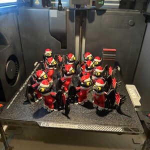 May include: A 3D printed model of a black dragon wearing a red Santa hat. The dragon is printed in multiple poses and is sitting on a black textured surface.