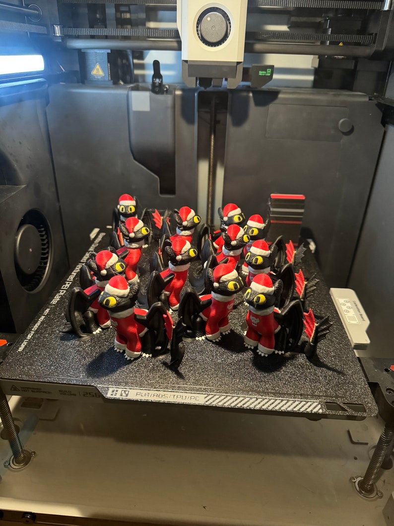 May include: A 3D printed model of a black dragon wearing a red Santa hat. The dragon is printed in multiple poses and is sitting on a black textured surface.