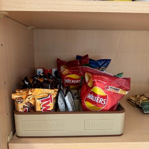 May include: A beige plastic bin labeled "CHOC BARS" and "SNACKS" holds various packaged snacks, including chocolate bars and bags of chips. The bin sits on a wooden shelf inside a cabinet, with tea boxes visible above.