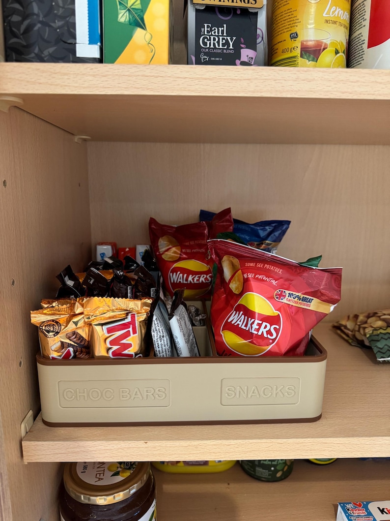 May include: A beige plastic bin labeled "CHOC BARS" and "SNACKS" holds various packaged snacks, including chocolate bars and bags of chips. The bin sits on a wooden shelf inside a cabinet, with tea boxes visible above.