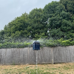May include: A blue, box-shaped object, resembling a police call box, is suspended in mid-air with water spraying around it. The background features a wooden fence, green foliage, and a cloudy sky. The object is a whimsical, creative piece.