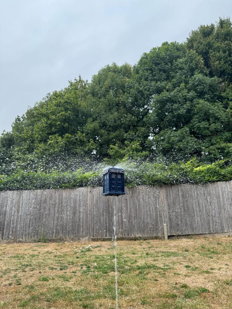 May include: A blue, box-shaped object, resembling a police call box, is suspended in mid-air with water spraying around it. The background features a wooden fence, green foliage, and a cloudy sky. The object is a whimsical, creative piece.