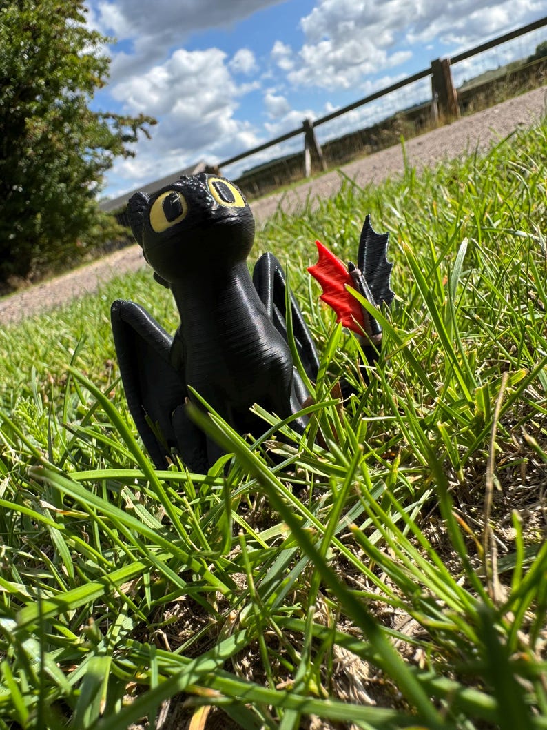 May include: A black dragon figurine with yellow eyes and red wing accents sits in green grass. The dragon is posed with its wings spread, and the background includes a fence, a path, and a blue sky with clouds.