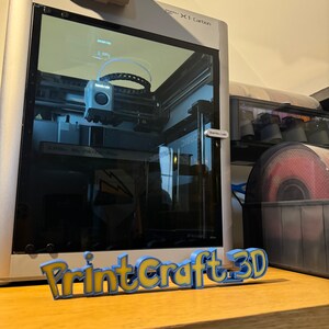 May include: A 3D printer with a clear plastic enclosure, showing the printing process in progress. The printer is labeled "X1-Carbon". A 3D printed sign reads "PrintCraft 3D".