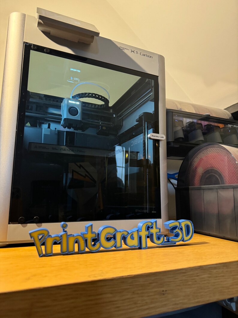 May include: A 3D printer with a clear plastic enclosure, showing the printing process in progress. The printer is labeled "X1-Carbon". A 3D printed sign reads "PrintCraft 3D".