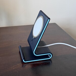 May include: A black phone stand with a light blue trim and a white charging pad. The stand is on a dark wooden surface. A white charging cable is connected to the stand.