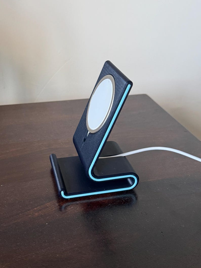 May include: A black phone stand with a light blue trim and a white charging pad. The stand is on a dark wooden surface. A white charging cable is connected to the stand.