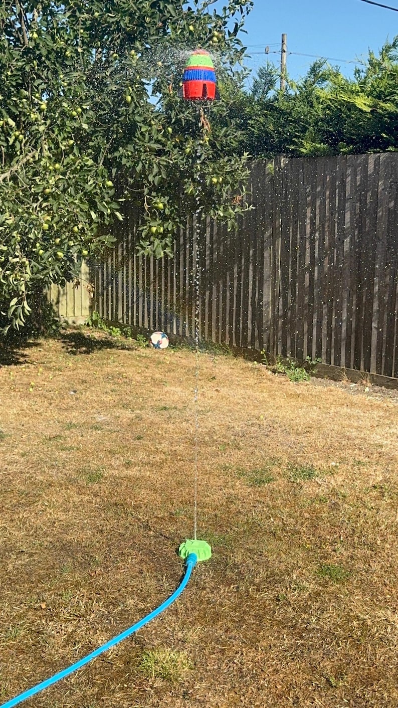 May include: A colorful pi&ntilde;ata hanging from a tree, spraying water. A blue hose connects to a green sprinkler on the ground. A soccer ball sits on the dry grass. The scene is outdoors on a sunny day.