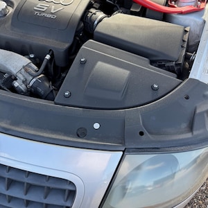 May include: A close-up view of a car engine bay. The engine cover is black with the Audi logo and "SV TURBO" text. A red bar spans the top. The front of the car is silver with a headlight and grill.