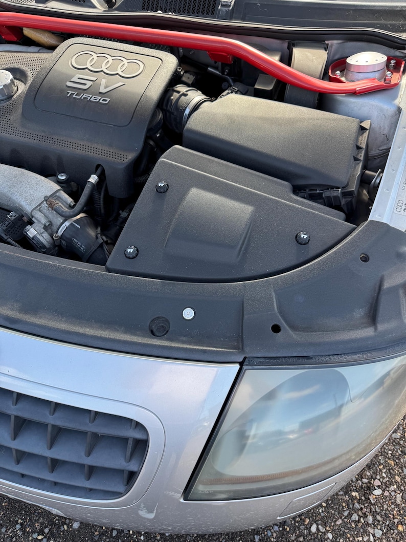 May include: A close-up view of a car engine bay. The engine cover is black with the Audi logo and "SV TURBO" text. A red bar spans the top. The front of the car is silver with a headlight and grill.