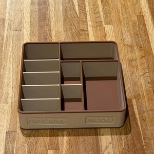 May include: A rectangular, two-tone beige and brown snack organizer with multiple compartments. The organizer has the words "CHOC BARS" and "SNACKS" printed on the front. The compartments are designed to hold various types of snacks.