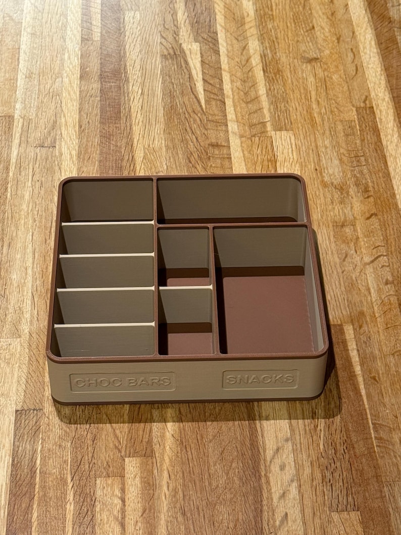 May include: A rectangular, two-tone beige and brown snack organizer with multiple compartments. The organizer has the words "CHOC BARS" and "SNACKS" printed on the front. The compartments are designed to hold various types of snacks.