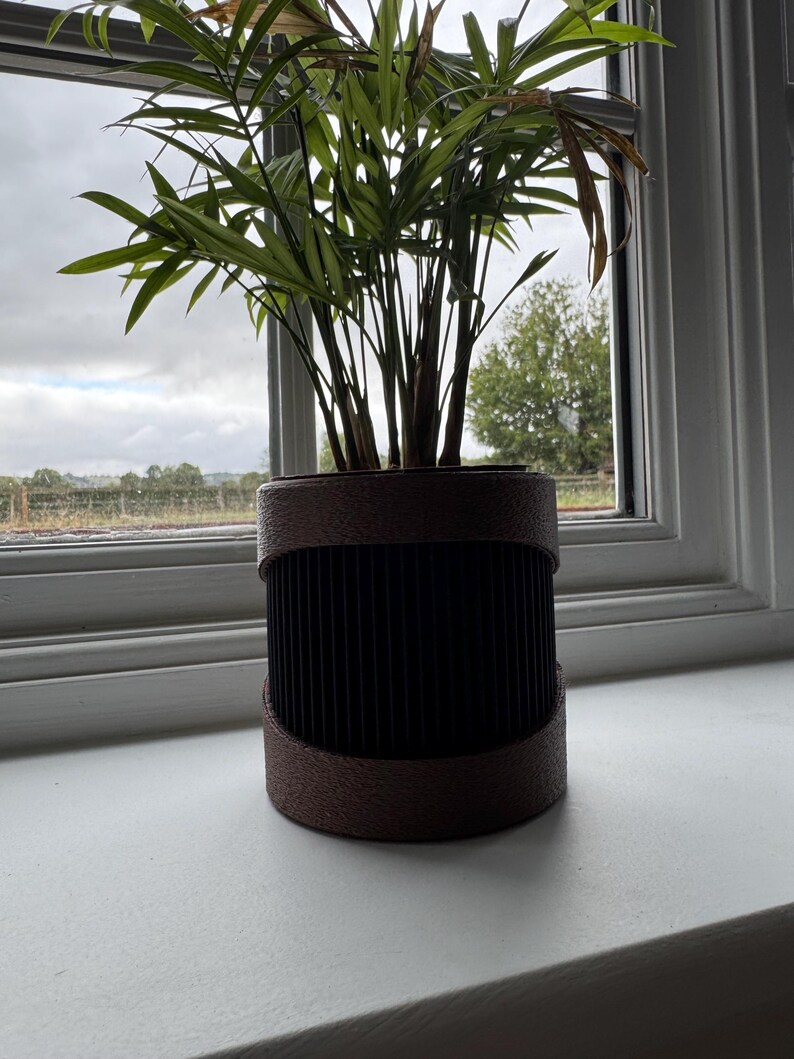May include: A potted plant with long, green leaves and brown stems sits in a decorative, cylindrical planter. The planter has a dark brown, ribbed center and a lighter brown rim. The plant is placed on a white windowsill.