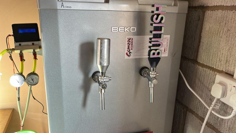 May include: A silver refrigerator with two beer taps, one labelled "Bullish". The refrigerator is labelled "BEKO" and has a digital display on the top left.
