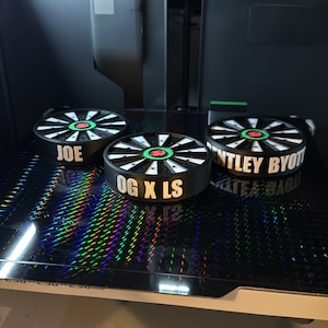 May include: Three black and white circular objects with text are displayed on a reflective surface. The objects have a dartboard design with green and red accents. The text on the objects reads "JOE", "OG X LS", and "BENTLEY BYOTT".