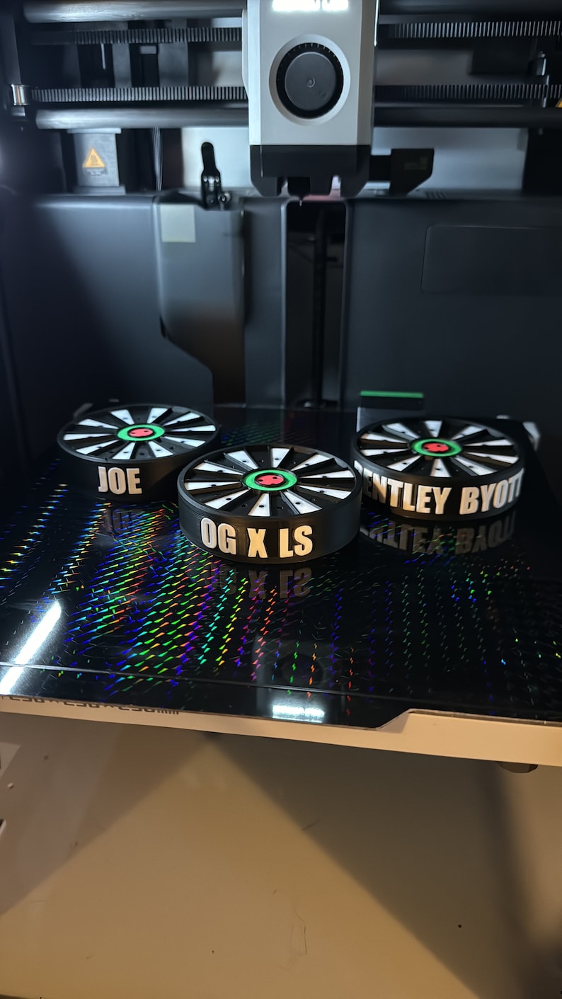 May include: Three black and white circular objects with text are displayed on a reflective surface. The objects have a dartboard design with green and red accents. The text on the objects reads "JOE", "OG X LS", and "BENTLEY BYOTT".