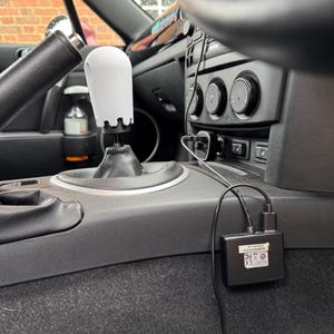 Op de afbeelding: Een witte, 3D-geprinte versnellingspookknop met een uniek ontwerp, ge&iuml;nstalleerd in een auto. Het interieur heeft een zwart dashboard, stuurwiel en middenconsole. Een zwart elektronisch apparaat met kabels is ook zichtbaar.