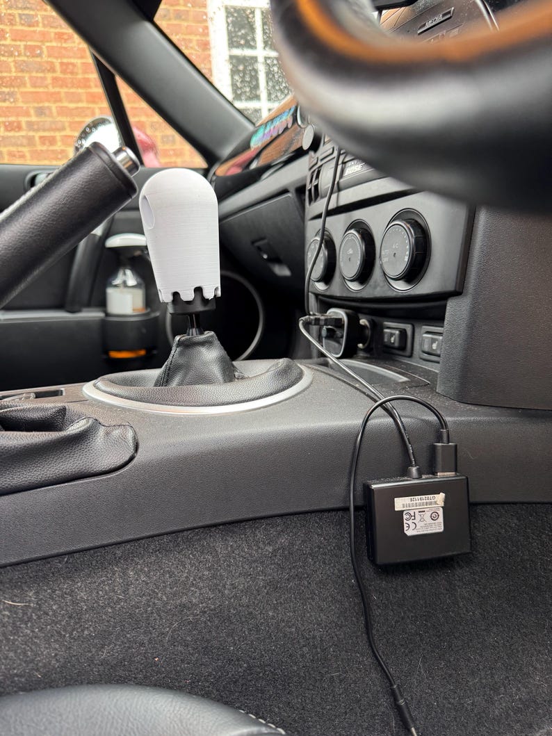 Op de afbeelding: Een witte, 3D-geprinte versnellingspookknop met een uniek ontwerp, ge&iuml;nstalleerd in een auto. Het interieur heeft een zwart dashboard, stuurwiel en middenconsole. Een zwart elektronisch apparaat met kabels is ook zichtbaar.